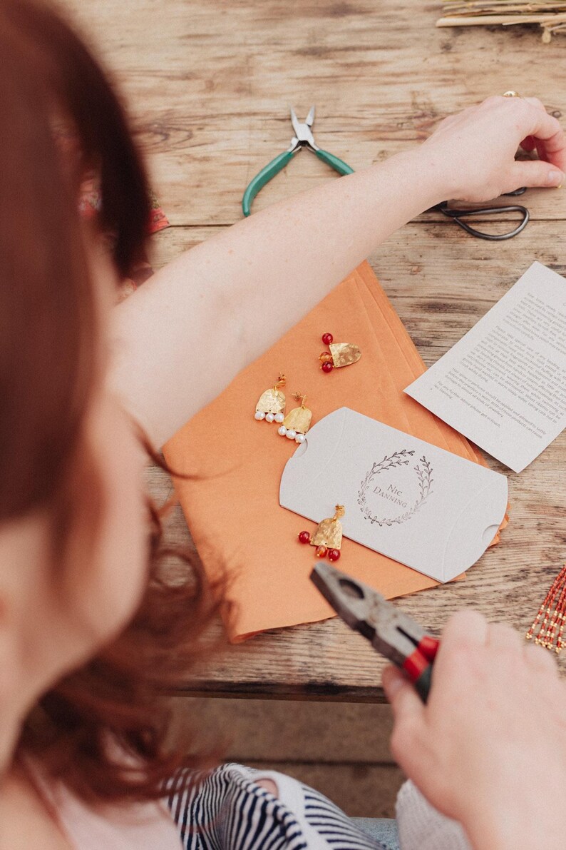 May include: Handmade gold earrings with white pearls and red beads are arranged on an orange surface. A small white box with a floral design and the text "Nic Darling" is also present. Pliers and other tools are visible, suggesting a craft setting.