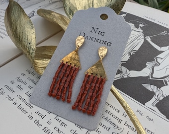 May include: A pair of gold earrings with red beaded fringe. The earrings are on a card with the text "Nic Danning".
