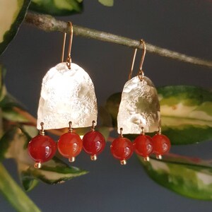 May include: Gold-tone earrings with a textured, arched top and three dangling red-orange beads. The earrings hang from gold-colored ear wires. The background is a blurred mix of green and gray.