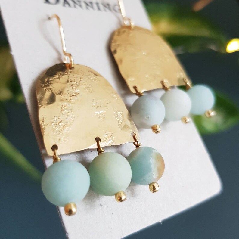 Pu&ograve; includere: Orecchini color oro con una texture martellata, con una parte superiore curva e tre perline pendenti verde-azzurro chiaro. Gli orecchini hanno una semplice chiusura a gancio e piccoli accenti dorati. Il design &egrave; minimalista ed elegante.