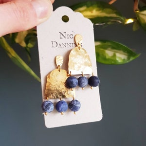 May include: Gold-tone earrings with a hammered texture and three dangling blue and gray beads. The earrings are attached to a cream-colored tag with the name "Nic Danning" printed on it.