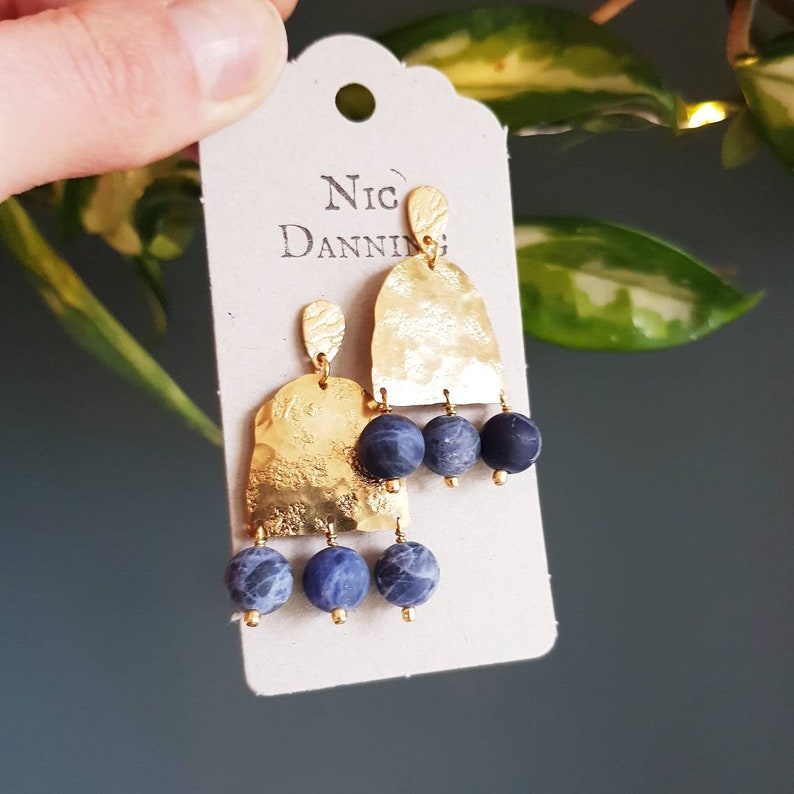 May include: Gold-tone earrings with a hammered texture and three dangling blue and gray beads. The earrings are attached to a cream-colored tag with the name "Nic Danning" printed on it.