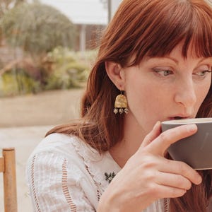 May include: A woman with red hair wearing gold earrings and rings, holding a gray mug. The earrings have a textured, bell-like shape with dangling beads. She is wearing a white blouse with embroidered details. The background is a blurred view of a window.
