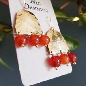 May include: Gold-tone earrings with a hammered texture, featuring three dangling orange-red beads. The earrings have a hook closure and are displayed on a white card with the text "Nic Danning".