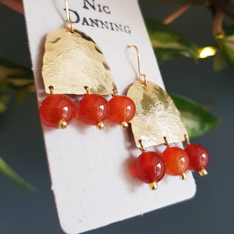May include: Gold-tone earrings with a hammered texture, featuring three dangling orange-red beads. The earrings have a hook closure and are displayed on a white card with the text "Nic Danning".