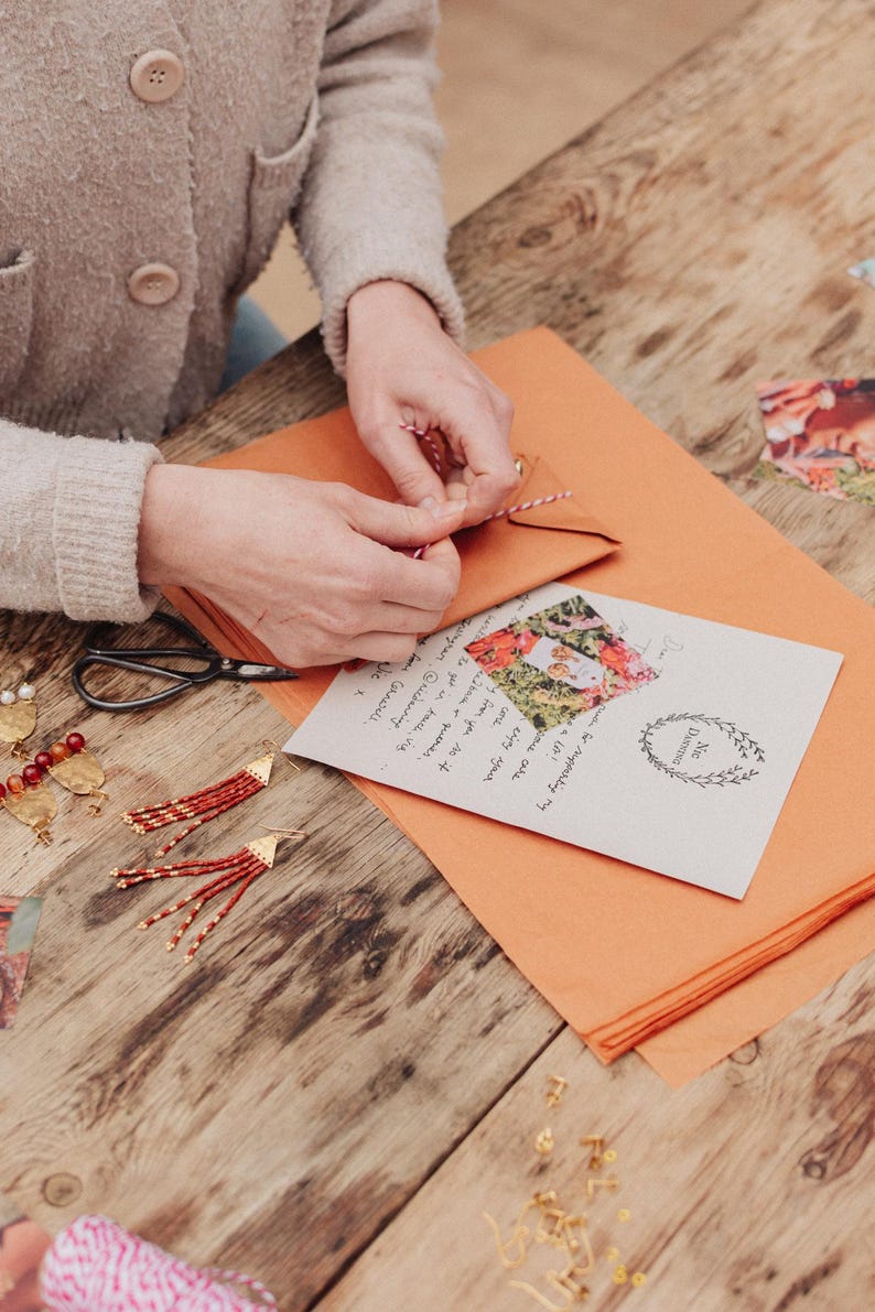 May include: A person is tying a red and white string around a small brown envelope. A greeting card with a floral photo and handwritten text is on an orange paper. Red and gold earrings and scissors are on a wooden table.