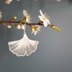 Puede incluir: Un pendiente plateado en forma de abanico con un diseño texturizado detallado, colgado de un gancho plateado. El pendiente cuelga de una rama con flores y capullos blancos, sobre un fondo gris apagado.