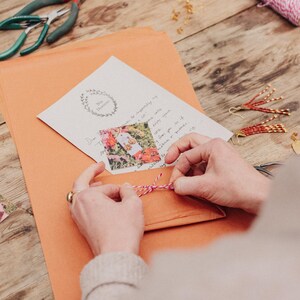 May include: An orange envelope is tied with pink and white twine. A handwritten note with a floral image is inside. Jewelry-making tools and finished earrings are on a wooden surface.