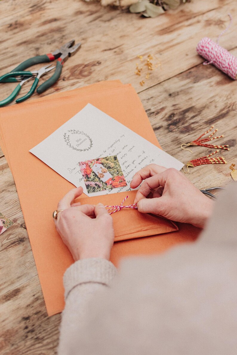 May include: An orange envelope is tied with pink and white twine. A handwritten note with a floral image is inside. Jewelry-making tools and finished earrings are on a wooden surface.