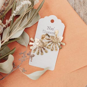 May include: A pair of gold-colored earrings with a floral design, displayed on a white tag that reads "Nic Danning". The earrings are set against an orange envelope and surrounded by dried flowers and greenery.