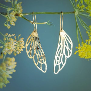 Może przedstawiać: Para złotych kolczyków w kształcie delikatnych skrzydeł motyla. Kolczyki wiszą na gałęzi z zielonymi liśćmi i żółtymi kwiatami.