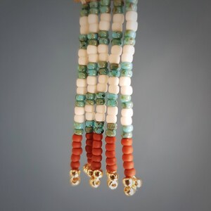 May include: A beaded tassel with white, turquoise, and brown beads. The tassel is made of multiple strands of beads and has gold accents at the bottom.