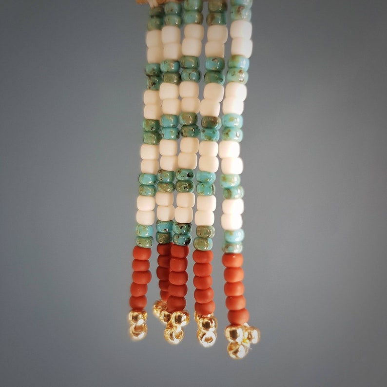 May include: A beaded tassel with white, turquoise, and brown beads. The tassel is made of multiple strands of beads and has gold accents at the bottom.
