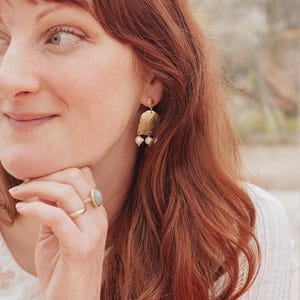 May include: Gold-toned earrings with a textured, rectangular design and three small, white pearls hanging from the bottom. A gold ring with a light green stone and a simple gold band are also visible.