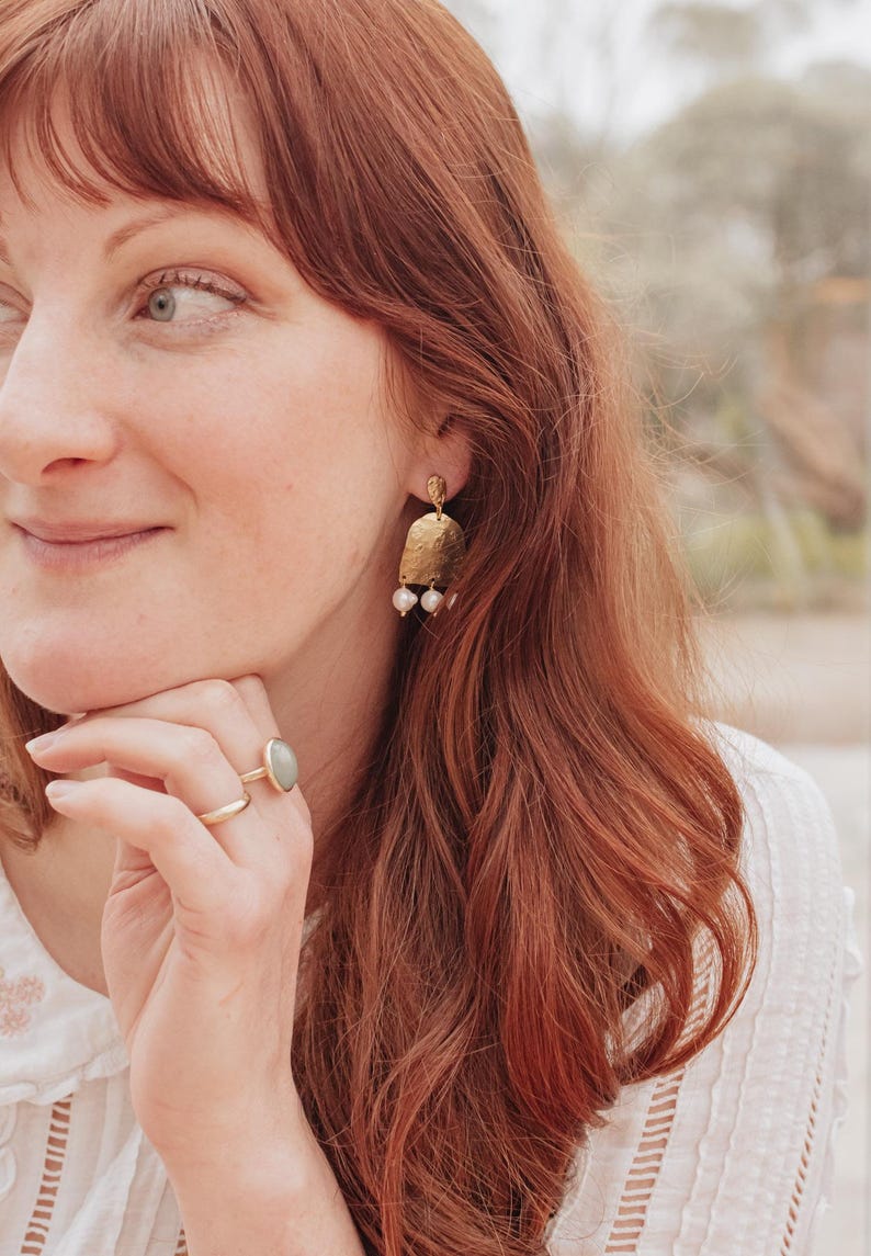 May include: Gold-toned earrings with a textured, rectangular design and three small, white pearls hanging from the bottom. A gold ring with a light green stone and a simple gold band are also visible.