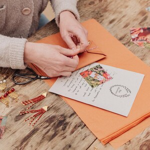 May include: A person is tying a red and white string around a small brown envelope. A greeting card with a floral photo and handwritten text is on an orange paper. Red and gold earrings and scissors are on a wooden table.