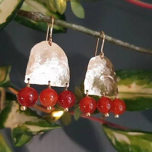 May include: A pair of gold-colored earrings with a hammered texture. Each earring features a half-moon shape with three red, round beads hanging below. The earrings are displayed against a backdrop of green foliage.