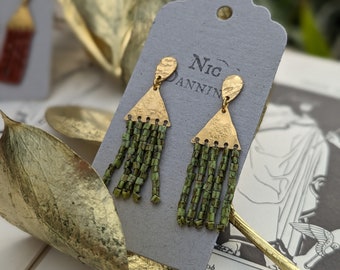 May include: A pair of gold dangle earrings with green beaded fringe. The earrings are on a white card with the text "NIC DANNING" printed on it.
