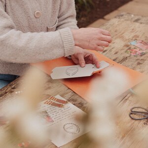 May include: A person working on a craft project, with hands holding a card with a wreath design. The scene includes an orange folder, scissors, and a wooden table. Other cards and stationery are also visible.