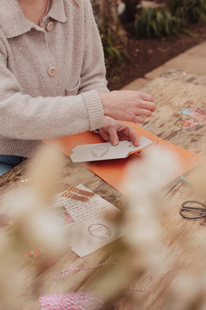 May include: A person working on a craft project, with hands holding a card with a wreath design. The scene includes an orange folder, scissors, and a wooden table. Other cards and stationery are also visible.
