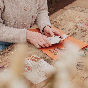 Pu&ograve; includere: Una persona tiene in mano un piccolo biglietto bianco con un disegno floreale, posto all'interno di una busta arancione. Altri articoli di cancelleria, tra cui una lettera con un bordo floreale, forbici e spago, sono su un tavolo di legno. La persona indossa un cardigan beige.