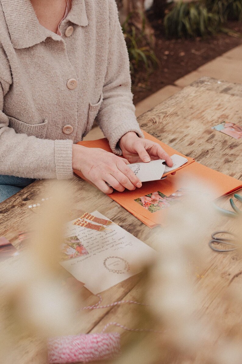 Pu&ograve; includere: Una persona tiene in mano un piccolo biglietto bianco con un disegno floreale, posto all'interno di una busta arancione. Altri articoli di cancelleria, tra cui una lettera con un bordo floreale, forbici e spago, sono su un tavolo di legno. La persona indossa un cardigan beige.