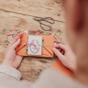 May include: An orange paper envelope tied with red and white twine, holding a small card with floral illustrations and the word "Dahlia". A pair of scissors and floral paper are on a wooden surface.