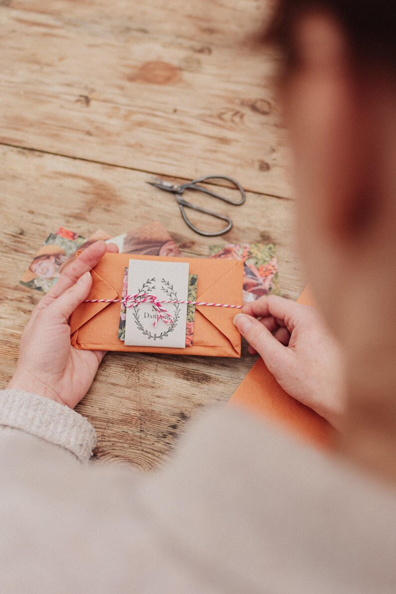 May include: An orange paper envelope tied with red and white twine, holding a small card with floral illustrations and the word "Dahlia". A pair of scissors and floral paper are on a wooden surface.