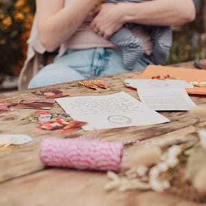 May include: A wooden table displays stationery items, including printed cards, envelopes, and a roll of pink and white twine. The scene is illuminated by natural light, suggesting a craft or stationery theme.
