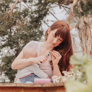 May include: A woman with red hair and a baby in her arms is using pliers to work on a craft project. A wooden table holds craft supplies, including pink and white twine and dried flowers. The scene is set outdoors with trees in the background.