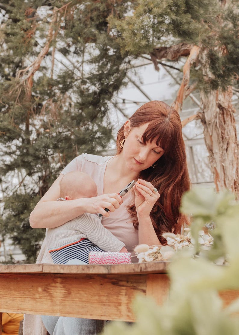 May include: A woman with red hair and a baby in her arms is using pliers to work on a craft project. A wooden table holds craft supplies, including pink and white twine and dried flowers. The scene is set outdoors with trees in the background.