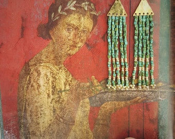 May include: A pair of long, beaded earrings with gold accents. The earrings are made of turquoise and gold beads and have a triangular shape at the top. The earrings are hanging in front of a painting of a woman.