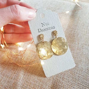 Può includere: Un paio di orecchini a disco color oro con una texture martellata, esposti su un'etichetta beige con il testo "Nic Danning". Gli orecchini sono tenuti da una mano, con luci di fata sullo sfondo.