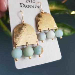 Pu&ograve; includere: Orecchini color oro con una texture martellata e tre perline verde-azzurro chiaro. Gli orecchini hanno una parte superiore curva e una piccola perla dorata alla base di ogni gemma. Gli orecchini sono esposti su una carta bianca con il testo "Nic Danning".