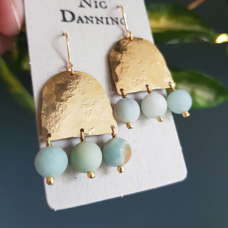 Pu&ograve; includere: Orecchini color oro con una texture martellata e tre perline verde-azzurro chiaro. Gli orecchini hanno una parte superiore curva e una piccola perla dorata alla base di ogni gemma. Gli orecchini sono esposti su una carta bianca con il testo "Nic Danning".