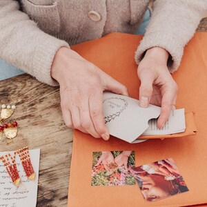 May include: A person is holding a small, light grey envelope with the text "Nic Danning" and a floral design. The envelope is inside an orange paper package. Earrings and a handwritten note are on a wooden surface.