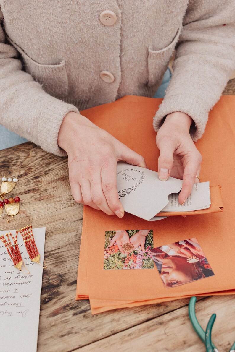 May include: A person is holding a small, light grey envelope with the text "Nic Danning" and a floral design. The envelope is inside an orange paper package. Earrings and a handwritten note are on a wooden surface.
