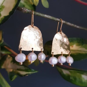 May include: A pair of gold-colored earrings with a hammered texture. Each earring features a curved, bell-shaped design with three dangling, pear-shaped white pearls. The earrings hang from gold-colored ear wires, showcasing a minimalist and elegant style.