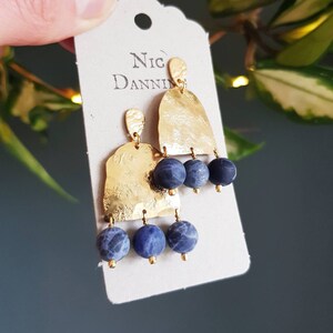 May include: Gold-tone earrings with a hammered texture and three dangling blue stone beads. The earrings are attached to a tag that reads "Nic Danning". The earrings are held up by a hand.