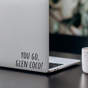 Puede incluir: Un portátil blanco con una pegatina negra que dice "You go, Glen Coco!" en la parte posterior. Una taza de cerámica blanca con motas marrones se encuentra sobre una mesa negra junto al portátil.