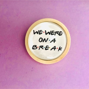 Puede incluir: Un posavasos redondo de madera con un centro blanco que presenta el texto "WE WERE ON A BREAK" en fuente manuscrita negra.