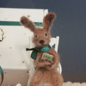 Può includere: Una statuetta di coniglio marrone e bianco seduta su una sedia bianca, che tiene un piccolo libro. Il coniglio indossa una sciarpa verde acqua. La statuetta è seduta su una superficie bianca con carta bianca sminuzzata.