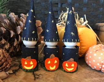 May include: Three small peg dolls wearing tall, dark blue felt hats decorated with yellow embroidered stars. Each doll features a cylindrical dark blue felt body adorned with a bright orange felt jack-o'-lantern face and green base trim.