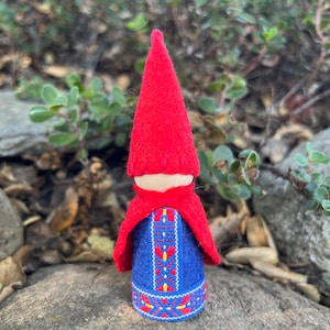 May include: A small felt gnome doll with a red pointed hat, a blue and red patterned dress, and a red cape.