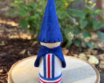 May include: A handmade gnome figurine with a blue felt hat and cape. The gnome's body is white with vertical red and blue stripes. The figurine is displayed on a wooden slice, with a blurred background of greenery and yellow flowers.
