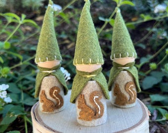 May include: Three small figurines with wooden heads and tall, pointed green felt hats. Each figure wears a green felt cape over a beige felt body, which features a brown felt squirrel appliqué with white embroidered details.