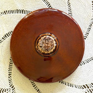 May include: A circular, glossy, dark brown ceramic lid featuring a centered, raised, textured knob with a mottled beige and brown finish. The lid has a smooth, reflective surface and a uniform, deep brown glaze throughout its entire top area.