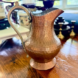 May include: A copper pitcher featuring a hammered texture, a wide pouring spout, and a curved metal handle. The vessel has a rounded body that tapers towards the neck and rests on a circular base, showcasing a metallic finish throughout.
