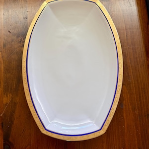 Peut inclure: Un plat de service ovale en porcelaine blanche doté d'un large bord doré avec un motif en relief complexe et d'une fine bande bleu cobalt vibrante bordant le bord intérieur de la garniture dorée, créant une présentation élégante.