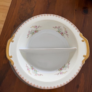 Pode incluir: Travessa de servir dividida creme com detalhes florais e acabamento dourado. A travessa tem duas alças e um divisor central. Padrões florais em rosa, amarelo e verde decoram a borda. Para petiscos.